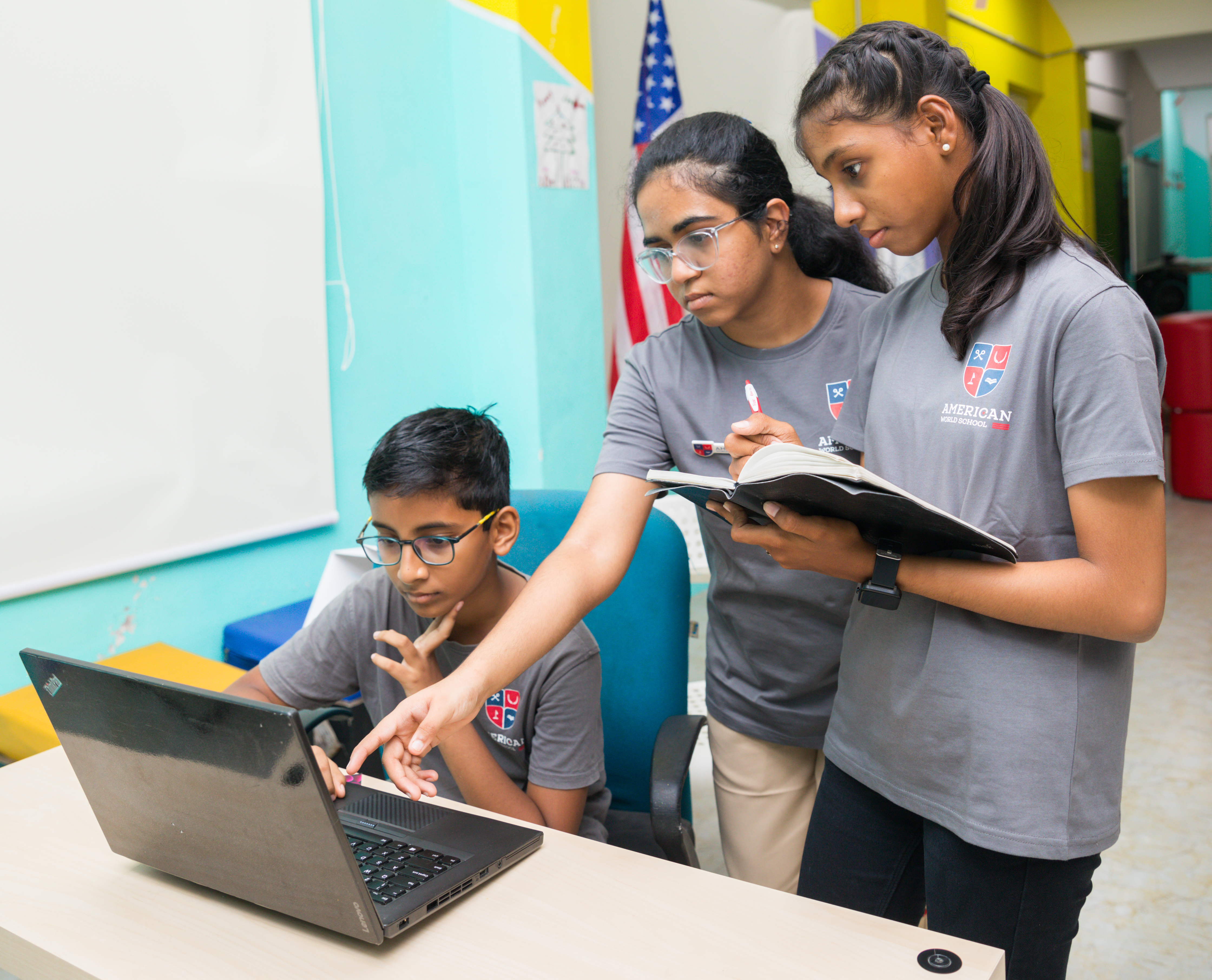 Two AWS girls coaching a younger student on a laptop, US flag behind